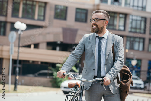 handsome businessman walking with bicycle on street in city and looking away