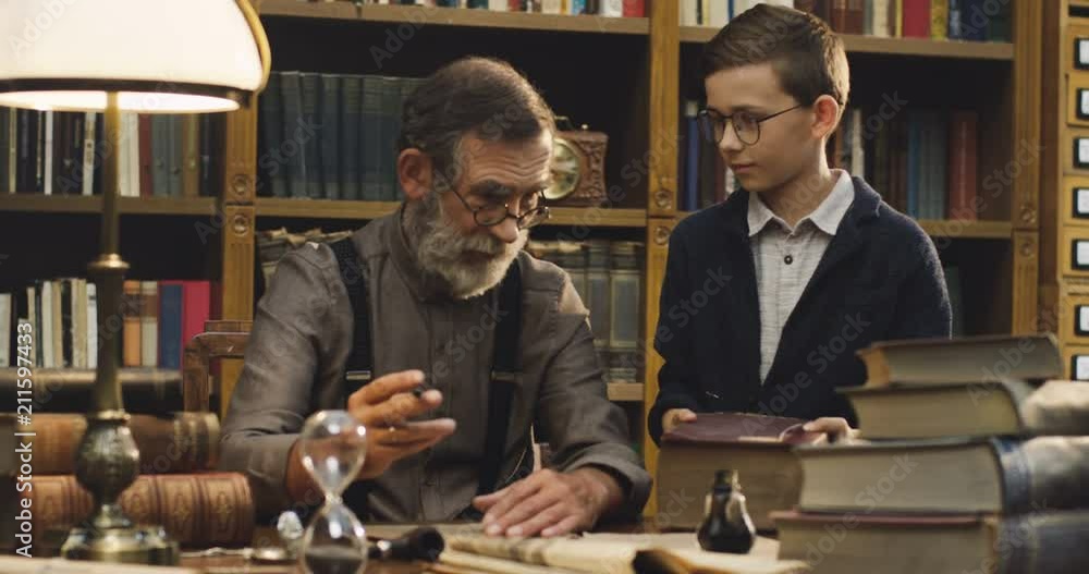 Old Caucasian man writing with an ink in the ancient library while his cute littile grandson bringing a big book on the table. Old Caucasian man writing with an ink in the ancient library while his cute littile grandson bringing a big book on the table.