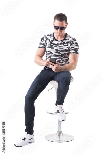 Handsome young adult man sitting on bar stool using and looking at cellphone. Full body isolated on white background. 