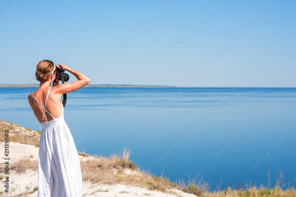 A girl in a white sarafan stands and takes pictures of the beautiful nature of rocks and water.