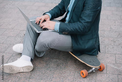 cropped view of man using laptop and sitting on skateboard
