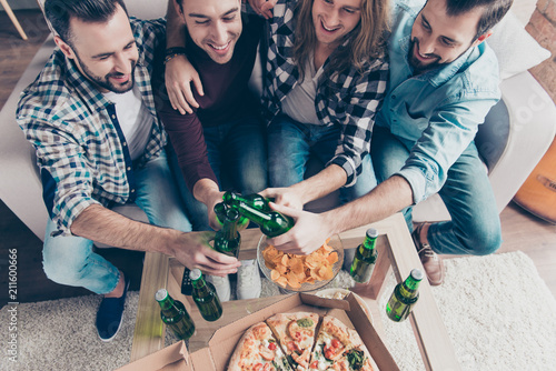 Cropped top view of four bearded, stylish, professional, successful, confident, attractive guys in casual, denim outfit clinking bottles with lager, having snacks on the table, group embracing