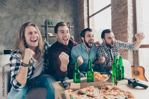 Four stylish, successful, attractive guys with modern hairstyle, raised fists, cheer for favorite team, watching hockey match, yelling, drinking lager eating chips, pizza, sitting in living room