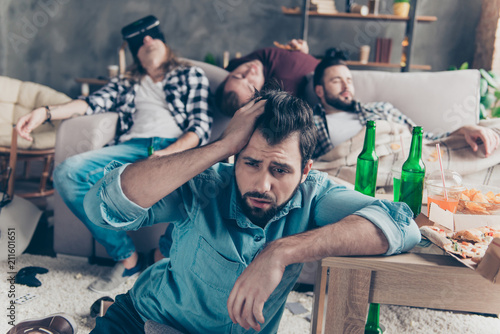 What we did last night? Bearded, upset, unhappy guy suffering from headache after night events, woke up on the floor holding hand on head, his drunk friends sleeping on couch on blurred background photo