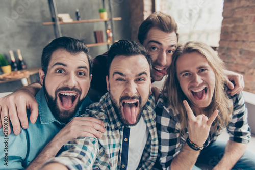 Self portrait of four attractive comic crazy funny foolish guys with hairstyle, showing tongue out and rock and roll symbol, making selfie on smart phone, front camera, sitting on couch in livingroom