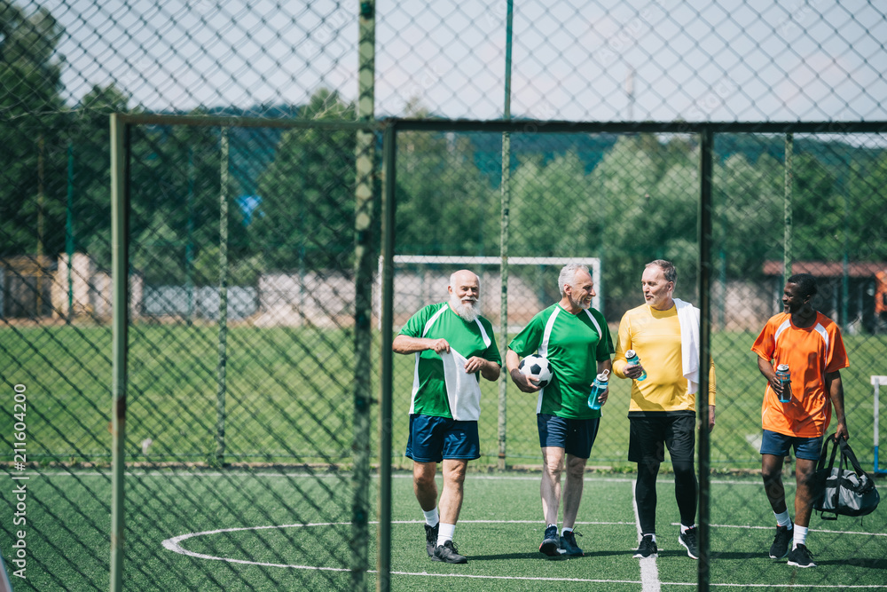 Obraz premium group of interracial elderly sportsmen with sportive water bottles walking on football field