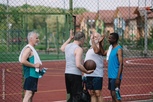 group of multiracial old sportsmen with basketball ball giving high five to each other on playground
