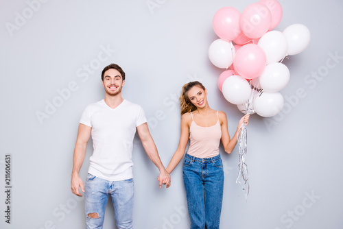 Portrait of attractive beautiful couple in casual outfits denim clothes holding pink and white air balloons holding hands looking at camera isolated on grey background