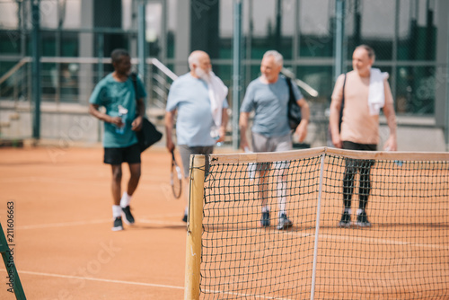 selective focus of multiracial elderly friends with tennis equipment on court © LIGHTFIELD STUDIOS