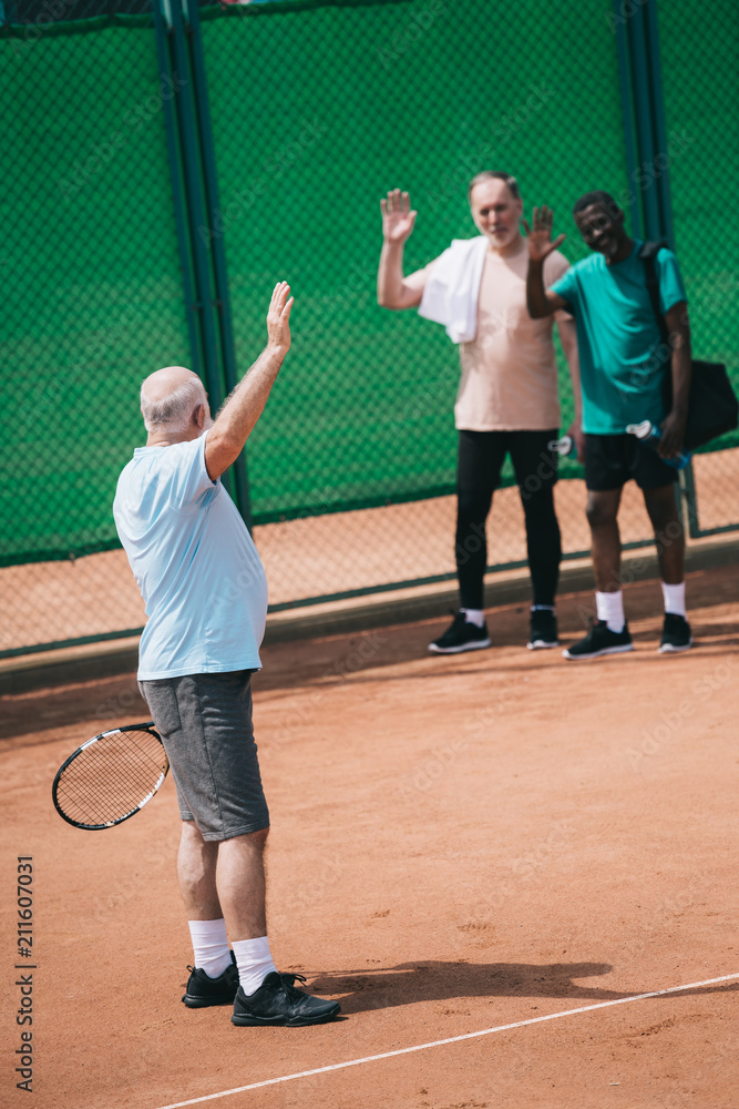 multiethnic old men greeting friend with tennis racquet on court