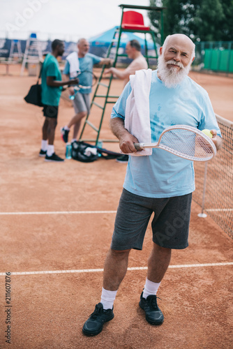 selective focus of elderly bearded man with towel and tennis racquet looking at camera while interracial friends having conversation on court © LIGHTFIELD STUDIOS