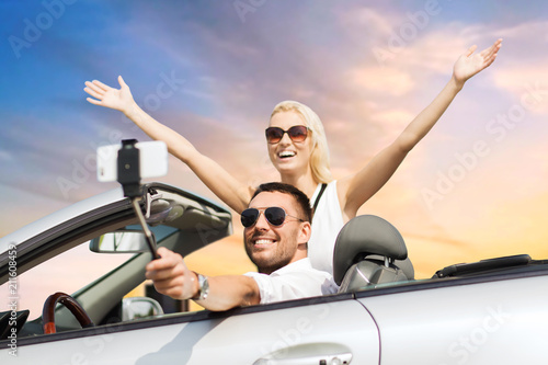 road trip, technology and travel concept - happy couple driving in convertible car and taking picture by smartphone on selfie stick over sky background © Syda Productions