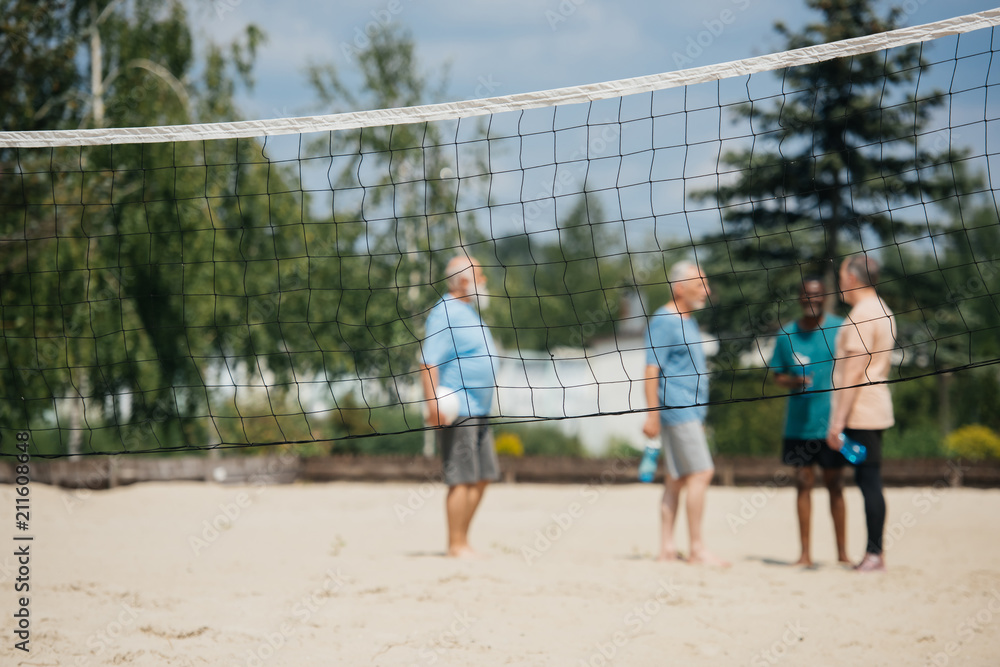 selective focus of net and interracial old men with volleyball ball on sandy beach