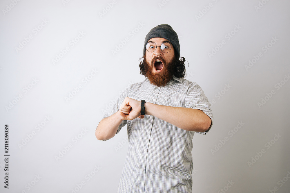 Suprised young bearded man wearing fur cap hat, glasses and white plaid shirt looking at the camera.