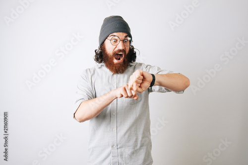 Surprised or scared young bearded hipster man wearing a fur hat cap and a plaid shirt and glasses looking at his hand watch.