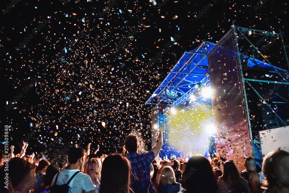 Huge concert crowd at night under the open sky on the roof. Pyrotechnic salute fireworks from multicolored red, blue and yellow confetti. Silhouettes of people having fun.