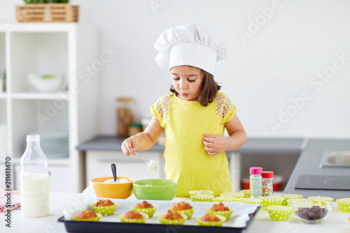 family, cooking, baking and people concept - little girl in chefs toque making batter for muffins or cupcakes at home kitchen