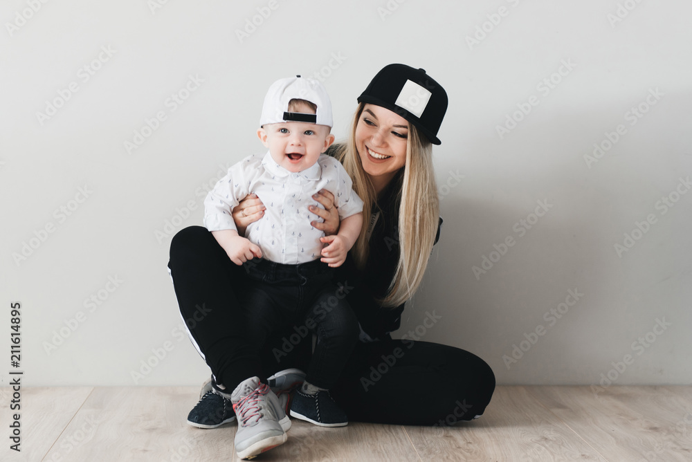 Stylish young mom in cap sitting and having fun with her cute one year old baby isolated on white background. Sports family