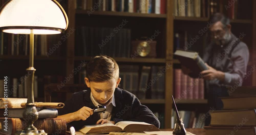 Little cute boy examining pictures in the book with a magnificent glass in hand while his grandpa looking at the book on the background in the ancient library. Little cute boy examining pictures in the book with a magnificent glass in hand while his grandpa looking at the book on the background in the ancient library.