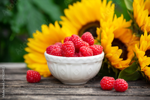 Ripe raspberries in white bowl with sunflower bouquet on wooden table, summer theme