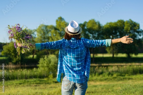 Outdoor summer portrait of woman with bouquet of wildflowers, straw hat. View from the back, female raised her hand.