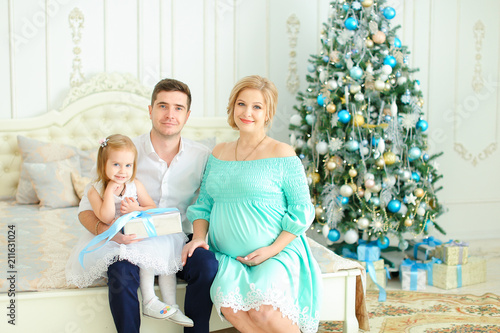 Little daughter sitting with happy father and pregnant mother on bed near decorated Christmas tree. Concept of european family celebrating winter holidays. photo