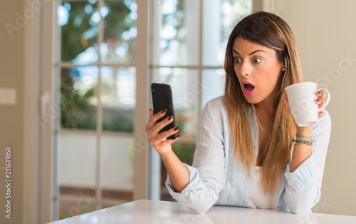 Fotografie Beautiful young woman looking at the phone suprised and excited while drinking cup of coffee at home