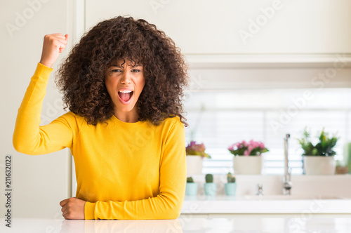 African american woman wearing yellow sweater at kitchen angry and mad raising fist frustrated and furious while shouting with anger. Rage and aggressive concept.