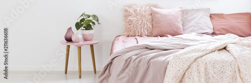 Real photo of a small table with a plant standing next to a bed with pink bedding in bedroom interior with white walls