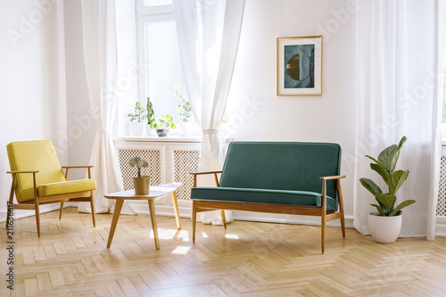 Wooden table between yellow armchair and green bench in white flat interior with poster. Real photo