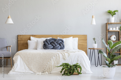 Blue knot pillow on bed with wooden headboard in grey bedroom interior with plants. Real photo