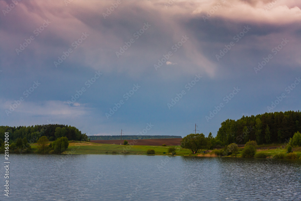 Evening colorful cloudy sky over a local lake and close to tiny woods. Lush undergrowth in the background and grassy field