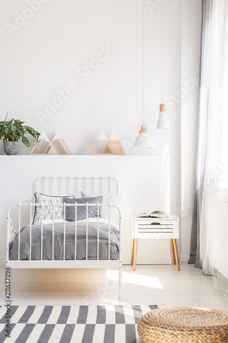 Pouf on patterned carpet near grey bed in white kid's bedroom interior with lamps. Real photo