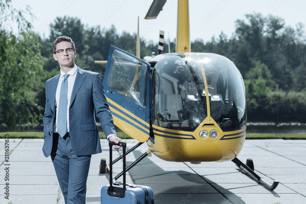 Personal belongings. Charming young CEO posing with his luggage while standing near a helicopter before the departure Personal belongings. Charming young CEO posing with his luggage while standing near a helicopter before the departure