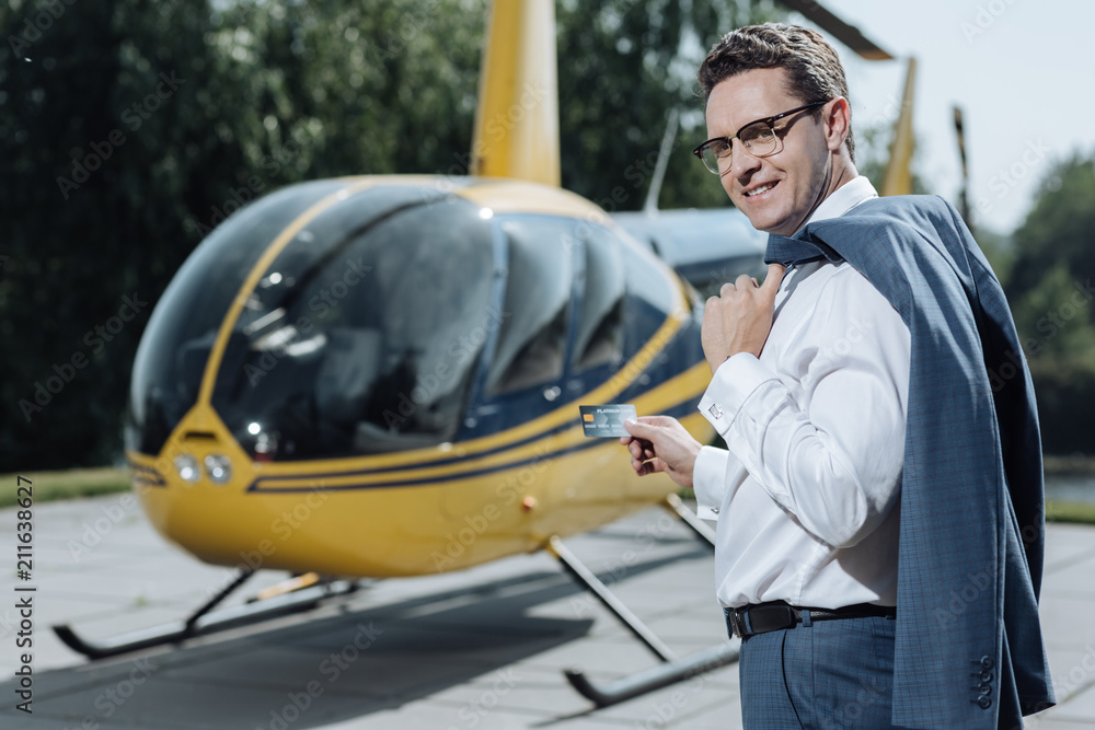 Perfect service. Cheerful young man posing with a credit card and smiling while standing at a helipad and being ready to pay for a rented helicopter Perfect service. Cheerful young man posing with a credit card and smiling while standing at a helipad and being ready to pay for a rented helicopter