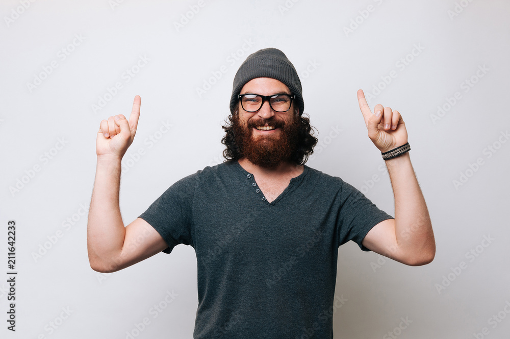 Naklejka premium Cheerful young bearded man wearing a T-shirt and glasses and a fur hat cap. Man pointing with hands and index fingers upwards. Offer concept.