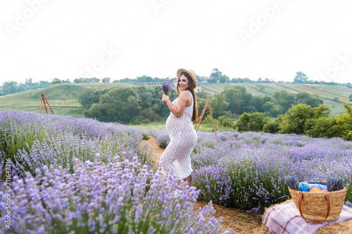 beautiful pregnant woman in white dress at violet lavender field with picnic basket on hay bale