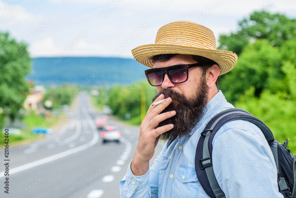 Old habit. Man with beard and mustache in straw hat smoking cigarette, road background defocused. Traveler stylish hipster take brake with cigarette. Smoking cigarette before long journey