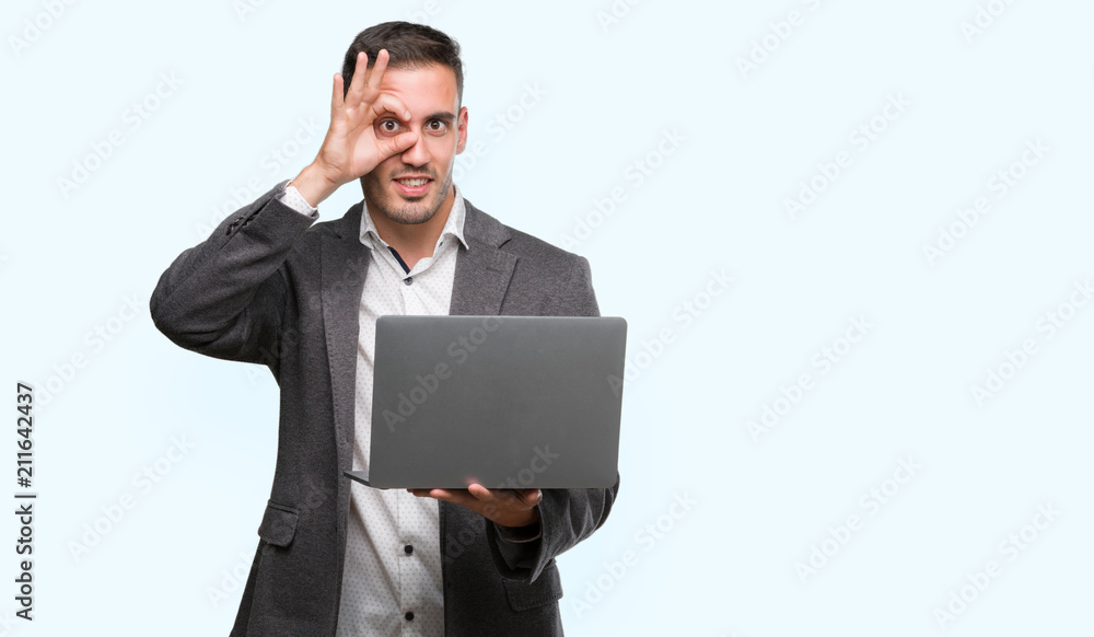 Handsome young man using computer laptop with happy face smiling doing ok sign with hand on eye looking through fingers Handsome young man using computer laptop with happy face smiling doing ok sign with hand on eye looking through fingers