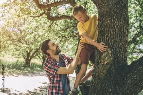 Bearded guy is standing near tree and supporting his son. Boy is climbing up. Child has put his hands on tree and looking at his father. They are smiling.