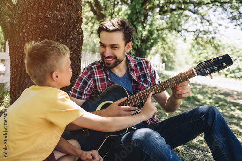 Small boy is leeaning to father's guitar. He wants to play on it. Bearded guy is holding this guitar and smiling to son. He looks happy. photo