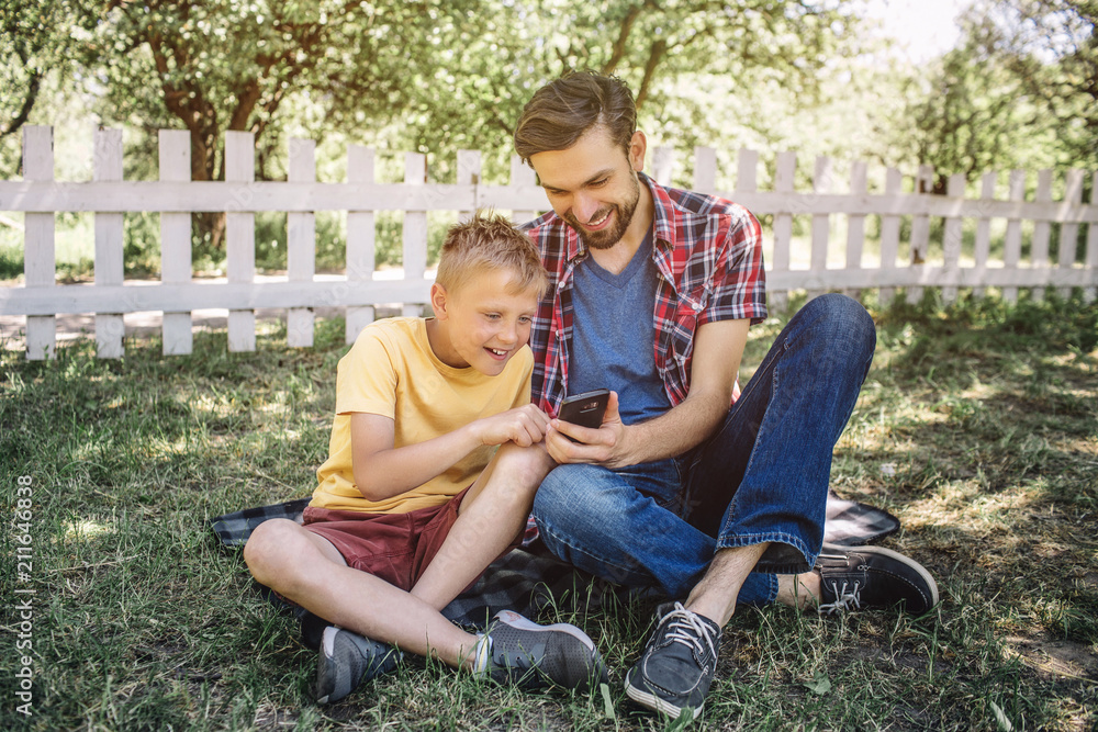 Man and child are sitting together on grass with their legs crossed and looking at phone with interest and wonder. THey are playing with it. Guys look happy and satisfied. Man and child are sitting together on grass with their legs crossed and looking at phone with interest and wonder. THey are playing with it. Guys look happy and satisfied.