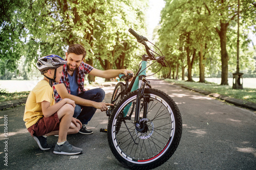 Guys have a short break during theit trip on bicycles. They are sitting at the border of road. Man is holding bicycle and looking at his son. Child is looking straight forward. photo