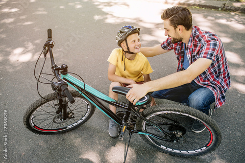 Nice and great picture of father and son sitting together in a squatt position and looking at each other. They are smiling. Man is hugging his son and holding bicycle at the same time. photo