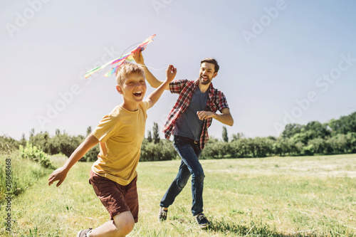 Happy kid is running on field. He is holding thread from kite. Father is holding kite. He is trying to run it to the sky. They are enjoying the moment.