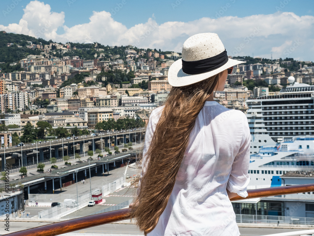 Beautiful, fashionable woman with long hair standing on the deck of a cruise ship against the background of a port with ships on a sunny, clear day. Concept of sea travel and recreation