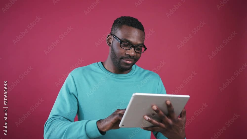 Young cute African student playing in video game on white digital tablet. Handsome guy wearing mint sweater smiling, looking at camera. Shiny white smile. Colored wall. Young cute African student playing in video game on white digital tablet. Handsome guy wearing mint sweater smiling, looking at camera. Shiny white smile. Colored wall.