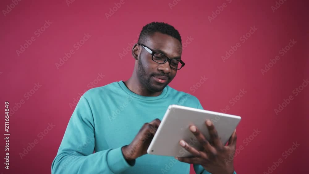 Portrait of attractive African guy in black spectacles wearing casual clothes. Young likable man holding digital tablet in hands, touching on background of colored wall. Portrait of attractive African guy in black spectacles wearing casual clothes. Young likable man holding digital tablet in hands, touching on background of colored wall.
