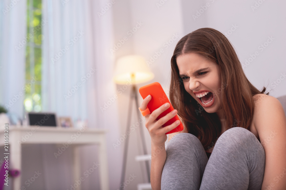 Unbelievable news. Low angle of wrathful teen girl shouting and holding phone for phone Unbelievable news. Low angle of wrathful teen girl shouting and holding phone for phone