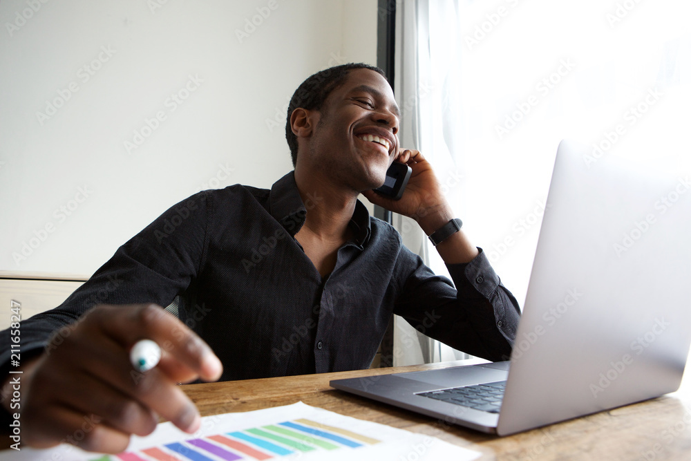 young african american business man sitting at desk with laptop talking on cellphone young african american business man sitting at desk with laptop talking on cellphone